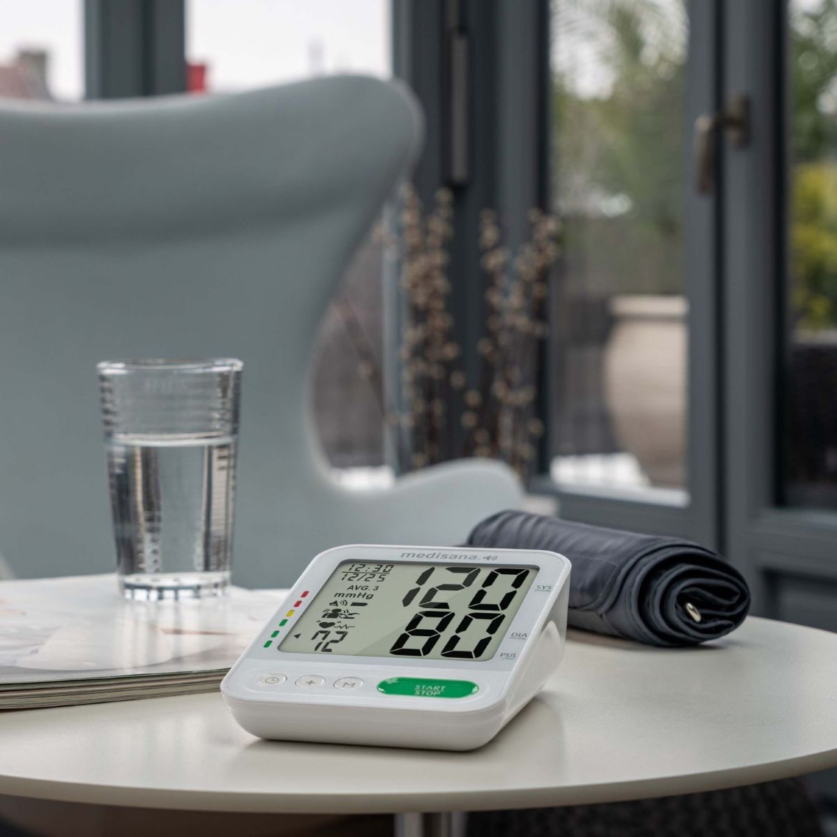 A digital blood pressure monitor sits ready for use on a small table, with the cuff placed beside it and a glass of water nearby.